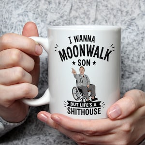 May include: White ceramic mug with the text "I WANNA MOONWALK SON BUT LIFE'S A SHITHOUSE" in black. The mug features an image of a man in a wheelchair pointing. The mug is held by a person.