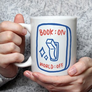 Könnte beinhalten: Weiße Keramiktasse mit blau-roter Grafik. Das Design zeigt ein blau umrandetes Rechteck mit den roten Worten "BOOK: ON" oben und "WORLD: OFF" unten. In der Mitte befindet sich eine blaue Schaltergrafik.