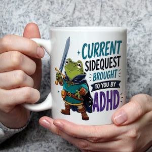 May include: White ceramic mug featuring a cartoon frog dressed as a knight, holding a sword. The mug has the text "CURRENT SIDEQUEST BROUGHT TO YOU BY ADHD" in teal and purple. The mug is held by a person.