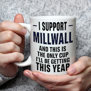 May include: A white ceramic mug featuring the dark blue text I SUPPORT MILLWALL and black text AND THIS IS THE ONLY CUP I'LL BE GETTING THIS YEAR printed on the front. The mug has a standard handle and a smooth, glossy finish.