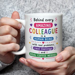 Op de afbeelding: Een witte keramische mok met kleurrijke tekst die luidt: Behind every AMAZING COLLEAGUE is a HUMAN BEING with real problems, real feelings and REAL COFFEE NEEDS! Het ontwerp bevat kleine paarse hartjes, gele sterren en roze accenten.