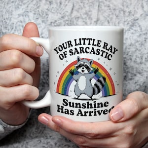 Pode incluir: Caneca de cerâmica branca com um desenho de guaxinim de desenho animado, um arco-íris e o texto "Your Little Ray of Sarcastic Sunshine Has Arrived". A caneca tem uma alça curva.