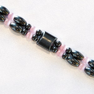 May include: A black and pink beaded bracelet with a magnetic clasp. The bracelet is made with small, round beads and larger, oval-shaped beads.