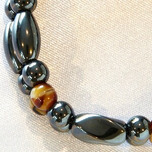 May include: A close-up of a bracelet made of black and brown beads. The bracelet is made of alternating black and brown beads, with the black beads being larger and more oval-shaped. The brown beads are smaller and rounder.