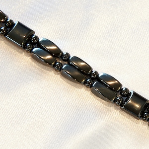 Op de afbeelding: Een zwarte hematiet armband met magnetische sluitingen. De armband is gemaakt van kleine, ronde kralen en rechthoekige hematiet schakels.