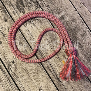 May include: A pink and gray rope with a tassel on one end. The rope is coiled and laid on a wooden surface.