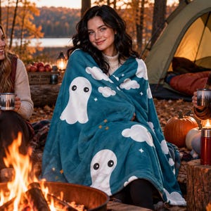 May include: A plush teal throw blanket featuring a repeating pattern of white ghosts, fluffy white clouds, and small orange stars. The soft fabric is draped over a person sitting outdoors near a campfire, showcasing the detailed ghost print.