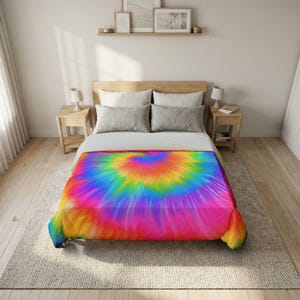 May include: A bedspread featuring a vibrant tie-dye pattern with a central spiral design in bright shades of pink, orange, yellow, green, and blue. The bedding is placed on a bed with two gray pillows and a white sheet set in a neutral bedroom.