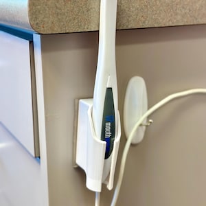 May include: A white dental intraoral scanner is held in a 3D-printed white holder. The scanner has a black and blue section with the word "mouth" printed on it. A white cord extends from the scanner.
