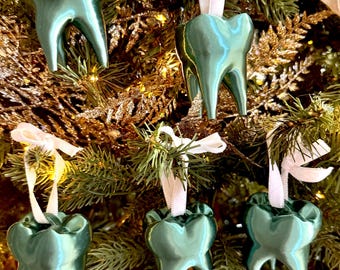 May include: Six teal-colored ceramic tooth ornaments hanging on a Christmas tree. Each tooth has a glossy finish and is suspended by a white ribbon. The tree has green needles and gold glitter accents.