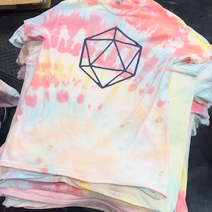 May include: A stack of tie-dye t-shirts in pastel colors, including pink, yellow, and blue. Each shirt features a dark blue geometric design. The shirts are made of soft material.