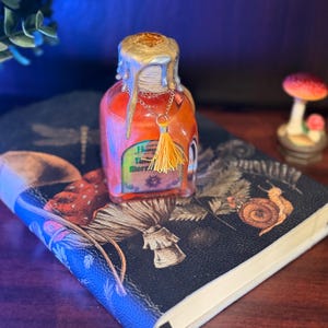 May include: A small, clear glass bottle filled with an orange liquid, topped with a gold-colored wax seal. A gold chain with a yellow tassel hangs from the bottle. The bottle rests on a dark book with illustrations of mushrooms and a snail.