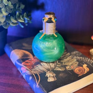 May include: A green, round glass bottle with a gold-colored top and a rope-wrapped neck. The bottle sits on a dark book with illustrations of mushrooms and a snail. The bottle's contents have a shimmering, swirling effect.