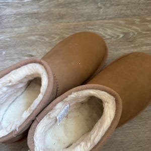 Könnte beinhalten: Ein Paar braune Wildlederstiefel mit weichem, weißem, flauschigem Innenfutter. Die Stiefel haben eine runde Spitze und eine kurze Knöchelhöhe. Die Außenseite ist einfarbig hellbraun.