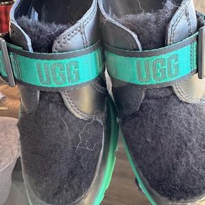 Puede incluir: Par de zapatos UGG negros con correa y suela verdes. La correa presenta el logotipo de UGG en verde. Los zapatos tienen un interior negro difuso y un exterior de cuero.