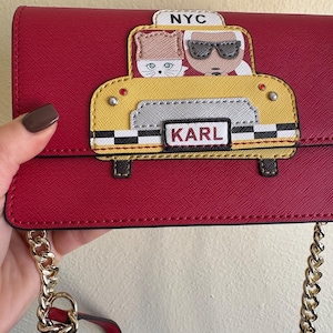 Puede incluir: Bolso bandolera rojo con un diseño de taxi amarillo con los textos "NYC" y "KARL". El bolso tiene una correa de cadena dorada y una correa roja. El diseño del taxi tiene dos personajes de dibujos animados.