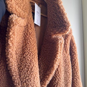 Può includere: Primo piano di un cappotto marrone in tessuto teddy con un esterno strutturato e morbido. Il cappotto ha un classico colletto a revers e una chiusura con bottoni. Il cappotto è appeso a una gruccia di legno.