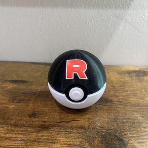 May include: A black and white Poke Ball with a red "R" printed on the top half.