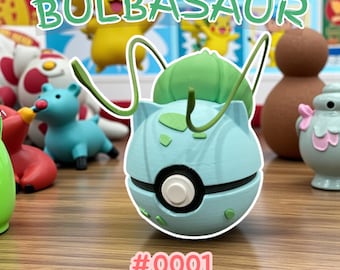 May include: A light blue Bulbasaur figure with a green bulb and vines. The figure has a black and white circle on the front and the number #0001 in pink. Other colourful figures are in the background.