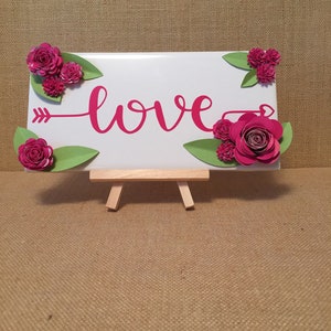 May include: A white sign with the word "love" written in pink cursive script. The sign is decorated with pink paper flowers and green leaves. The sign is on a small wooden easel.