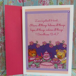 Puede incluir: Una tarjeta rosa y morada con una cita sobre el amor de 1 Corintios 13:4,7. La tarjeta presenta una ilustración de dos osos de peluche tomando el té.