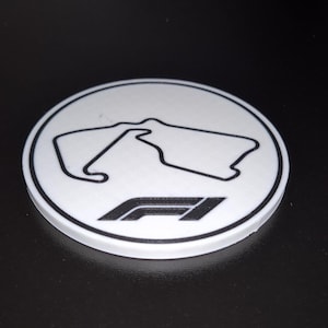 Könnte beinhalten: Ein weißer, kreisförmiger Untersetzer mit einer schwarzen Umrisszeichnung einer Rennstrecke und dem F1-Logo. Der Untersetzer liegt auf einer schwarzen Oberfläche. Das Design ist schlicht und modern, für jeden Formel-1-Fan.