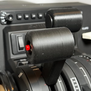 May include: Close-up of a black aircraft throttle control with a red indicator light. The throttle has a black lever and a round knob with a textured surface. The control is labelled "Flight Detent" and "Trim".