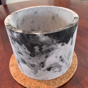 May include: A white and black marbled concrete planter with a cork coaster underneath. The planter is cylindrical and has a smooth, polished finish.