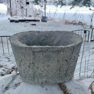 May include: A gray, textured concrete planter with a wide, open top. The planter has a rough, weathered appearance and is set against a snowy outdoor backdrop. The planter is designed for holding plants and flowers.