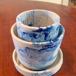Peut inclure: Ensemble de trois pots de fleurs en céramique blanche avec un motif bleu marbré. Les pots sont empilés, avec une petite soucoupe à la base. Les tourbillons bleus créent un aspect artistique unique, idéal pour les plantes d'intérieur.
