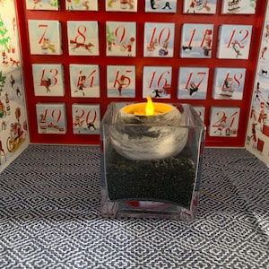 May include: A red Advent calendar with 24 numbered boxes, each featuring a festive illustration. A decorative candle with a faux flame sits in a clear glass container filled with black stones, placed on a blue and white patterned surface.