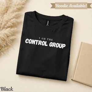 以下が含まれることがあります： 前面に白い文字で「I AM THE CONTROL GROUP」と書かれた黒いTシャツ。 シャツはきちんと折りたたまれています。 左下には「Black」の文字があります。 右側にはベージュの本があります。