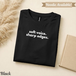 以下が含まれることがあります： 「soft voice. sharp edges.」の白文字がプリントされた黒いTシャツ。Tシャツは折りたたまれ、ニュートラルな背景に表示されています。「Hoodie Available!」の文字が上部のバナーにあります。