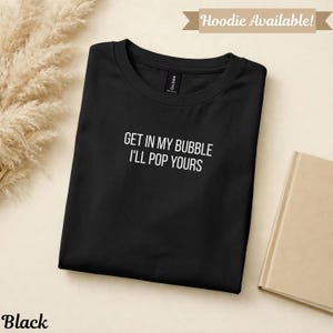 May include: Black t-shirt with the text "GET IN MY BUBBLE I'LL POP YOURS" in white. The shirt is folded and displayed on a neutral background. The word "Black" is at the bottom left.