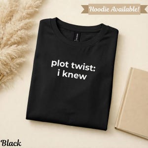 Peut inclure: T-shirt noir plié avec l'inscription blanche "plot twist: i knew". L'étiquette Gildan est visible. Un livre beige est en arrière-plan. La bannière en haut indique "Hoodie Available!".