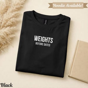 May include: Black t-shirt with the text "WEIGHTS BEFORE DATES" in white. The shirt is folded and displayed on a neutral background. A banner at the top reads "Hoodie Available!"