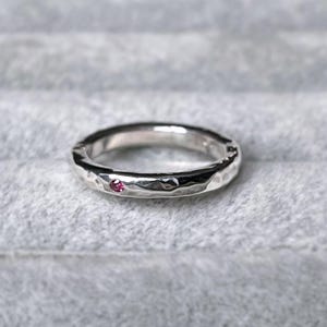 May include: A silver-toned ring with a textured, hammered surface finish. The band features a single, small, round, vibrant pink gemstone set flush into the metal, creating a subtle point of colour against the metallic silver band.