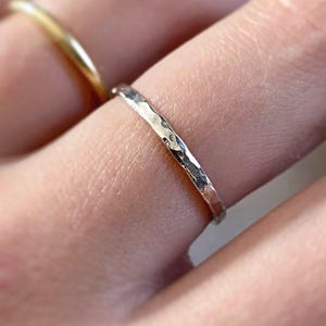 May include: A thin, silver-coloured ring with a hammered, textured surface worn on a finger. The ring features a reflective, metallic finish with irregular indentations across the band, creating a distinct, light-catching appearance on the metal.