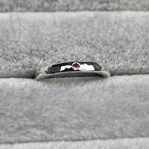 May include: A silver ring with a faceted band and a small, round, pink gemstone. The ring is set against a grey, textured background.