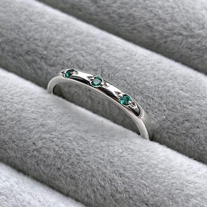 May include: A silver-toned metal ring featuring three small, round, vibrant green gemstones set flush into the band. The band has a smooth, polished finish and a slender profile, showcasing a clean design with the stones spaced evenly across the top.