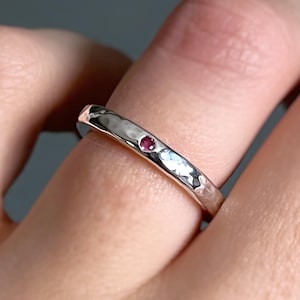 May include: A silver ring with a hammered texture and a small, round, red gemstone. The ring is shown on a finger, with a focus on the jewelry's design and details. The ring is a piece of jewellery.