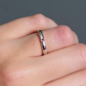 May include: A silver ring with a hammered texture and a small, round, red gemstone. The ring has a minimalist design. The gemstone is set in a bezel setting. The ring is a simple and elegant piece of jewellery.