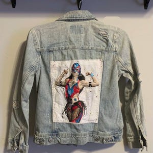 May include: A light-wash denim jacket featuring a large rectangular patch on the back. The patch displays an embroidered figure wearing a blue and red luchador mask and matching wrestling attire, posing with flexed arms. The jacket has distressed details.