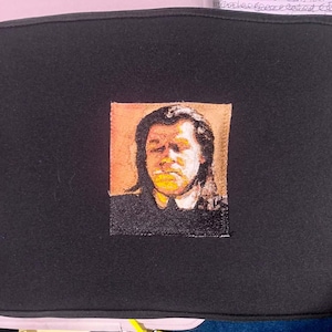 May include: A black rectangular laptop sleeve featuring a centered square patch with a portrait of a person with dark hair. The portrait is rendered in shades of orange, brown, and black. The sleeve has a zipper closure on the left side.