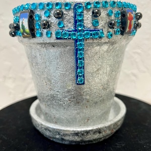 May include: Silver-colored flower pot with a matching saucer. The pot is decorated with a blue jeweled cross and a band of blue jewels and black beads around the rim. Small pictures are set into the rim.