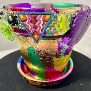 以下が含まれることがあります： 手描きの小さな植木鉢と受け皿。 金箔、紫、緑、ピンクの塗料で装飾されています。 三角形のデザイン、紫色の葉、装飾的な装飾が特徴です。
