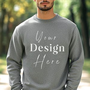 May include: A heather gray sweatshirt with the text "Your Design Here" in white script. The crew neck sweatshirt is a solid color and has long sleeves. The image is taken outdoors with a blurred background.
