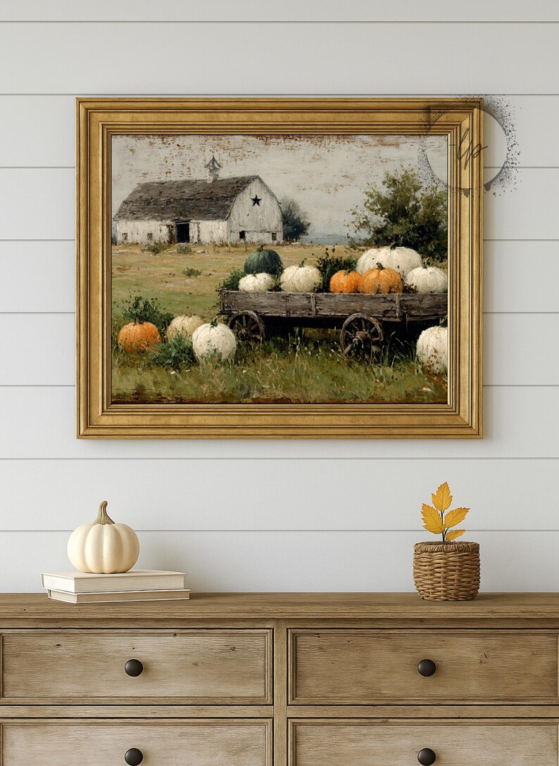 Op de afbeelding: Een ingelijste print met een rustieke herfstsc&egrave;ne. Een schuur met een ster op de achtergrond en een houten kar vol pompoenen in wit, oranje en groen. De print zit in een gouden lijst.
