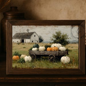 Op de afbeelding: Een ingelijste prent toont een rustieke herfstsc&egrave;ne. Een houten kar is gevuld met pompoenen in wit, oranje en groen. Een witte schuur met een ster staat op de achtergrond, in een veld.