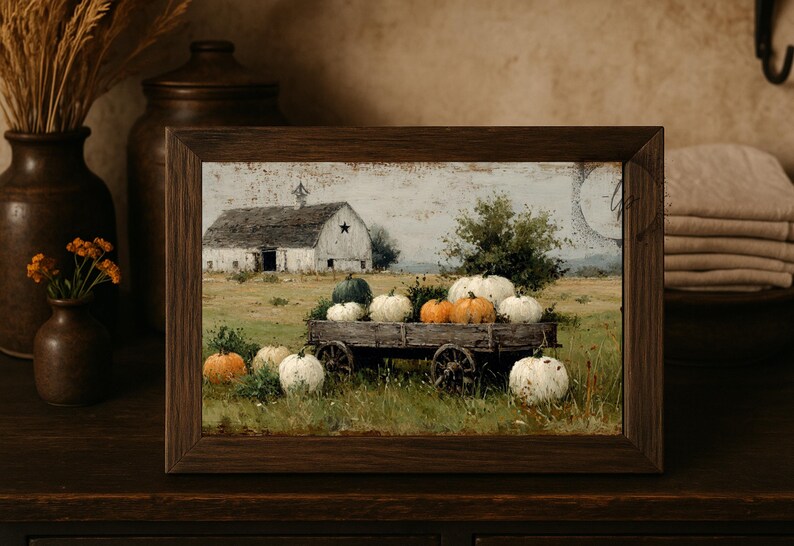 Op de afbeelding: Een ingelijste prent toont een rustieke herfstsc&egrave;ne. Een houten kar is gevuld met pompoenen in wit, oranje en groen. Een witte schuur met een ster staat op de achtergrond, in een veld.